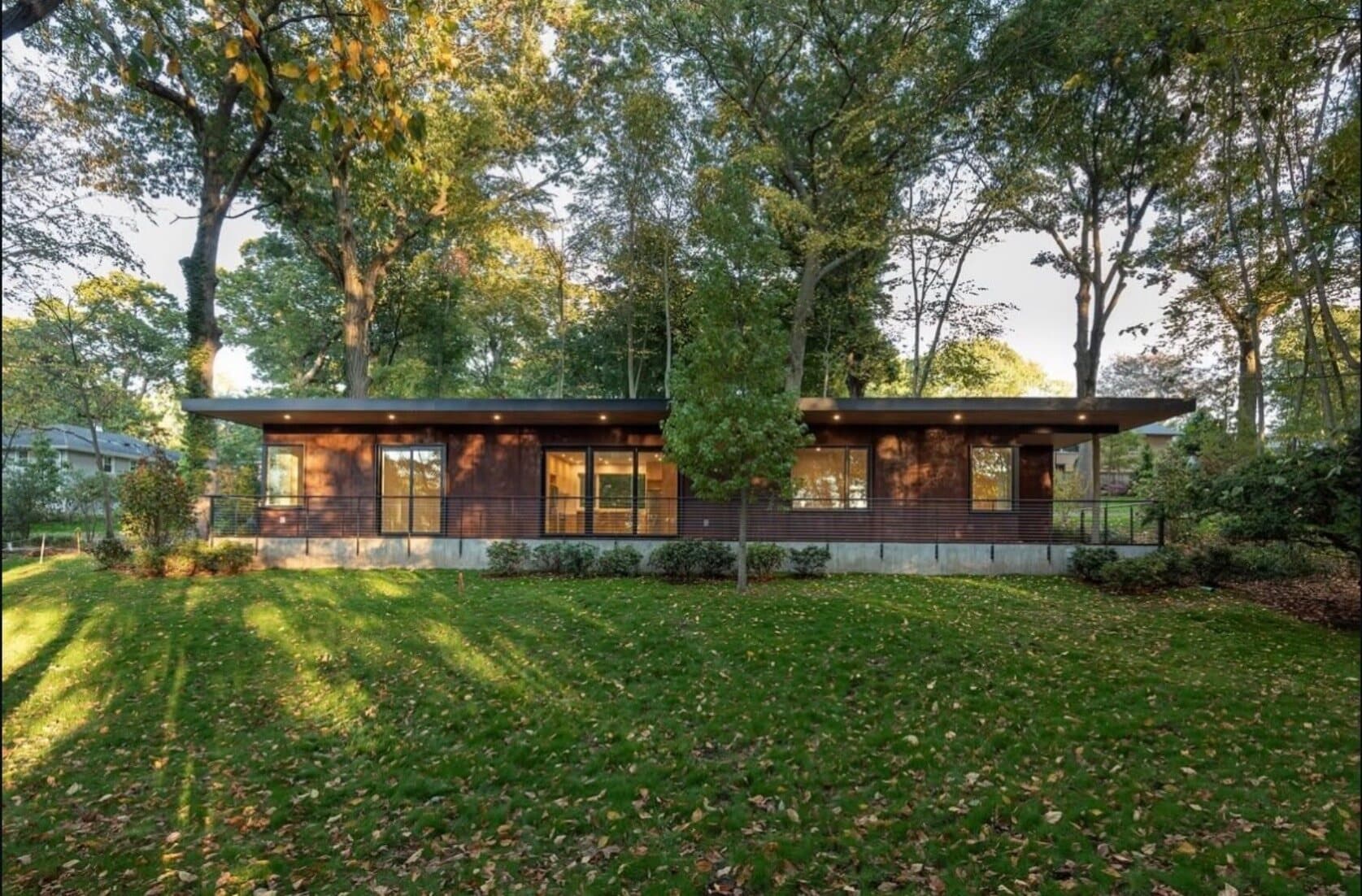 Photograph of the completed Yale Building Project house exterior at dusk, dark wood siding beneath a mature tree canopy, interior lights glowing through full-height glazing.