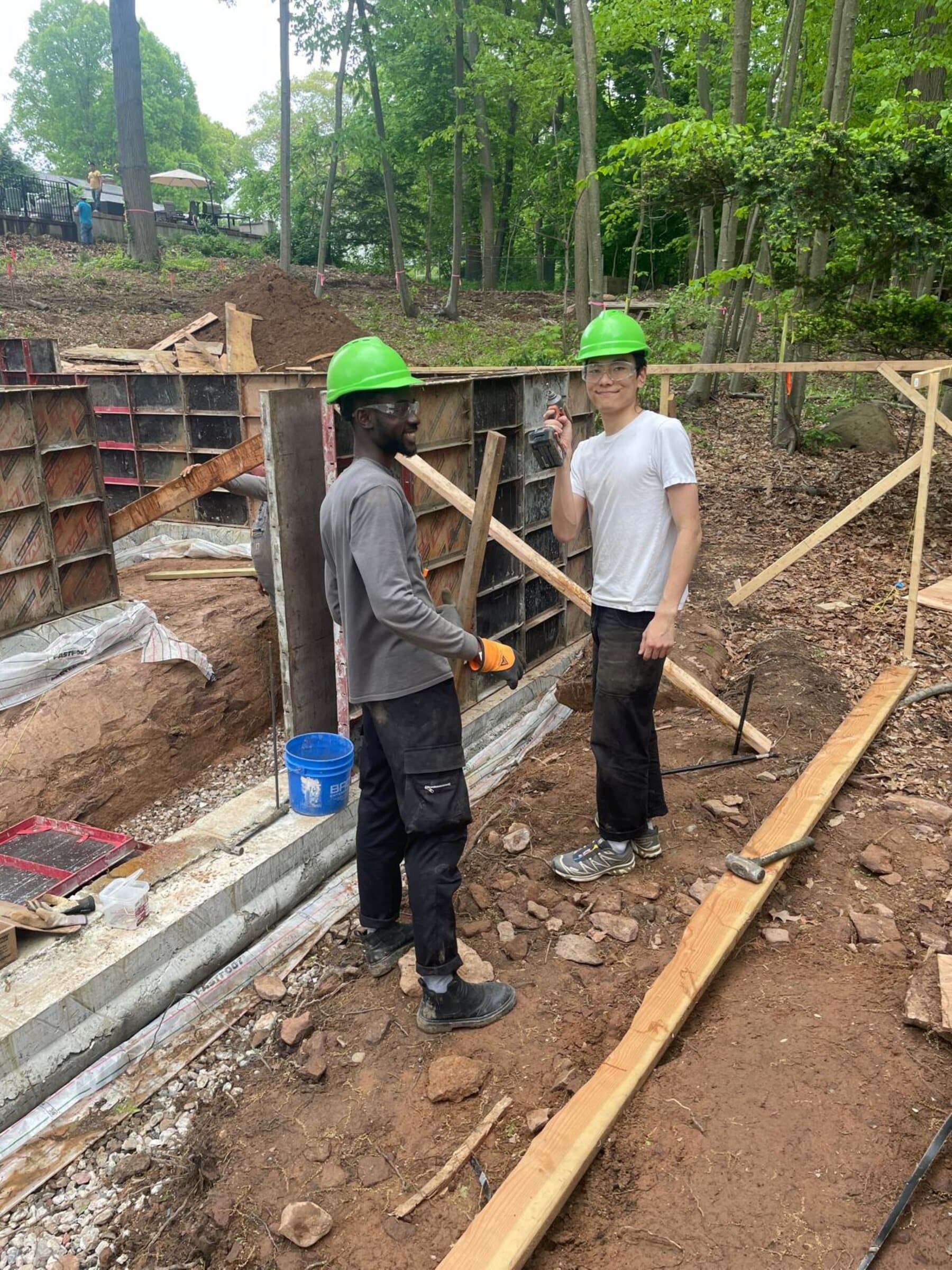 Construction photograph of two team members setting out foundation formwork on site, wearing green hard hats on excavated ground.