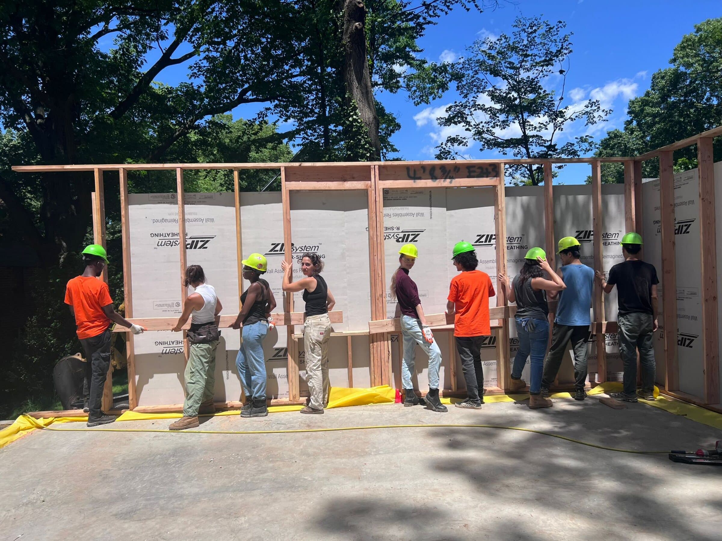 Construction photograph of the full team raising a framed timber wall wrapped in sheathing, wearing green hard hats.
