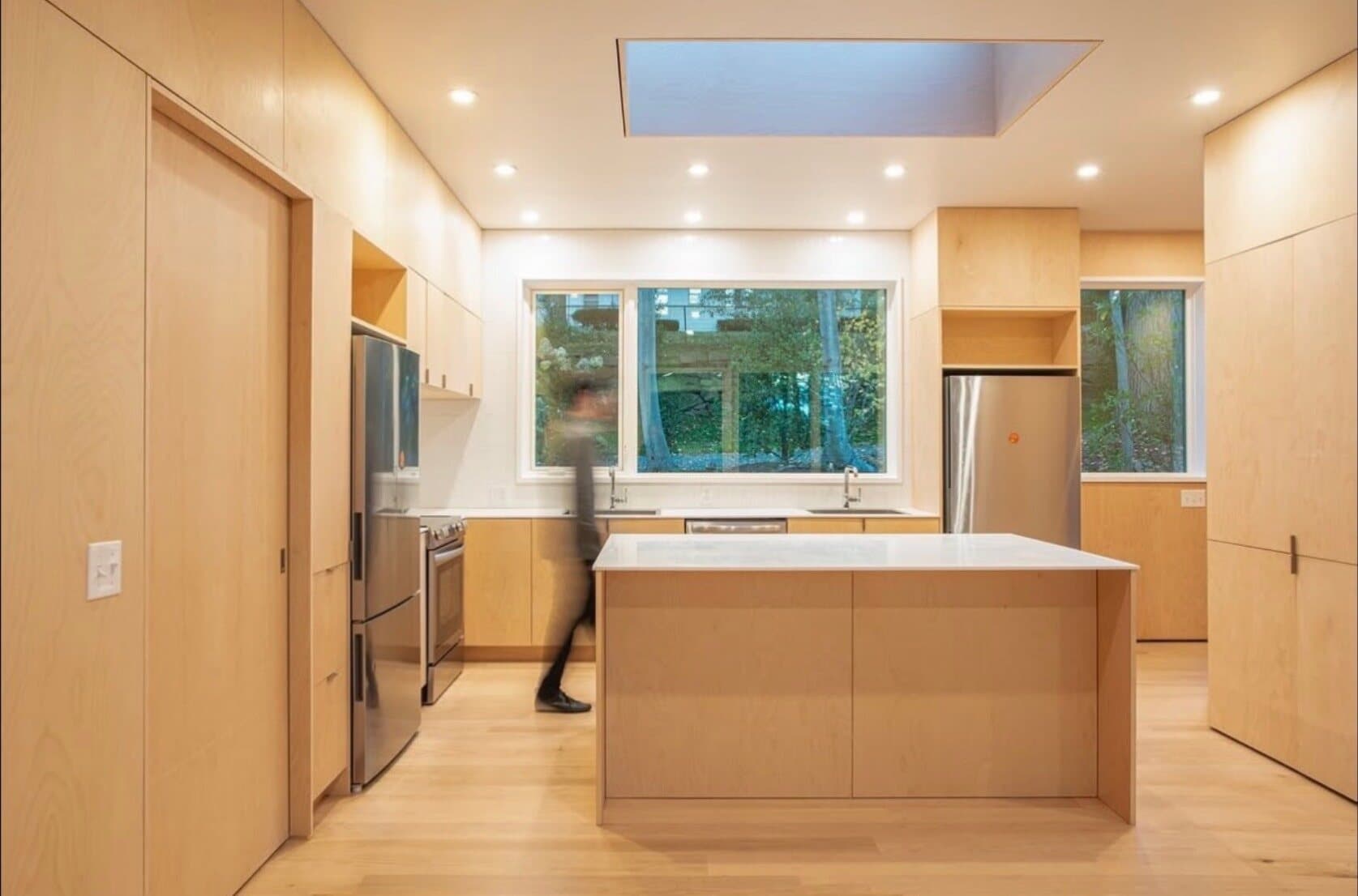Photograph of the finished house's shared kitchen, maple cabinetry, a skylight, and a figure walking past the island.